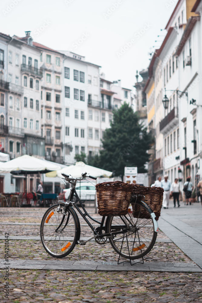 Charming scene in Coimbra: a bicycle with twin wicker baskets parked on a square under a cloudy morning sky. Old buildings with a tree, a terrace and people strolling add life to the quaint setting.