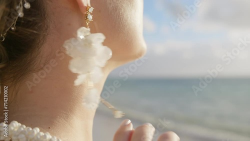Close up side view of beautiful woman wearing luxury hanging white flower earrings. Side view of bride slowly touching boho style floral drop earrings. Girls hand with wedding diamond ring at beach 4K photo