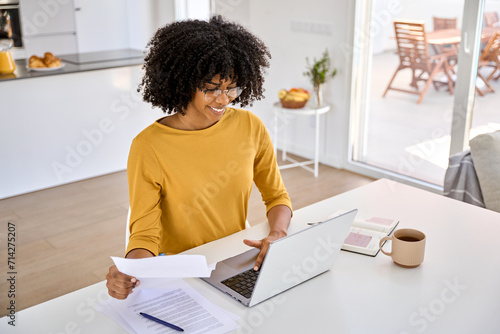 Young smiling happy African American woman checking financial paper calculating banking loan or household payments using laptop computer paying bills online sitting at home table in kitchen.