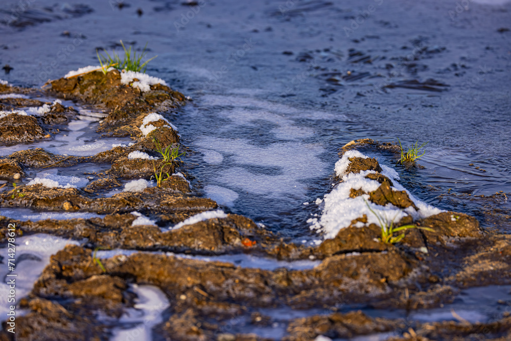 Blue ice of a puddle next to dirt from clods of earth of an agricultural field