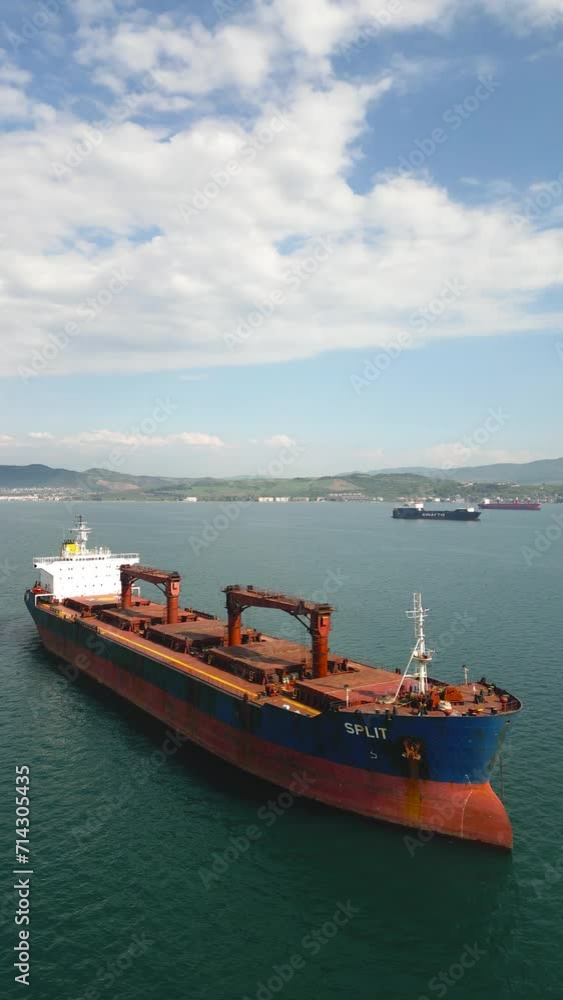 Aerial side view of red cargo carrier ship on the sea. Camera is sliding right to left. Residential area and clouds are visible. Aerial side view of red cargo carrier ship on the sea. Camera is sliding right to left. Residential area and clouds are visible.