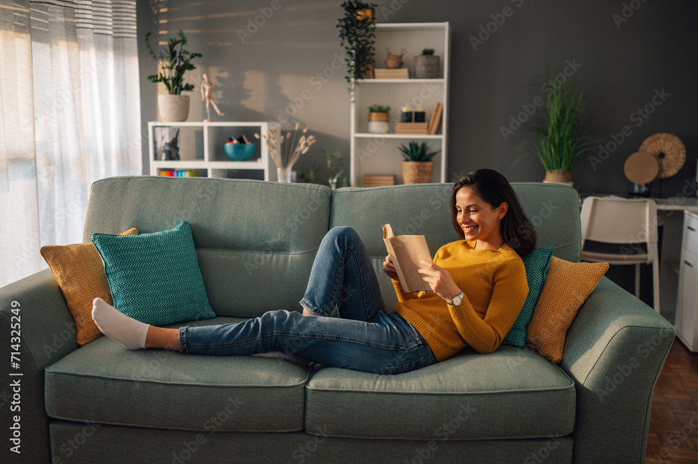 Obraz premium Young woman lying on comfortable sofa reading a book in her free time at home