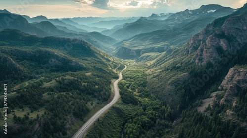  an aerial view of a mountain valley with a winding road in the foreground and trees on the other side of the valley, with a mountain range in the background.