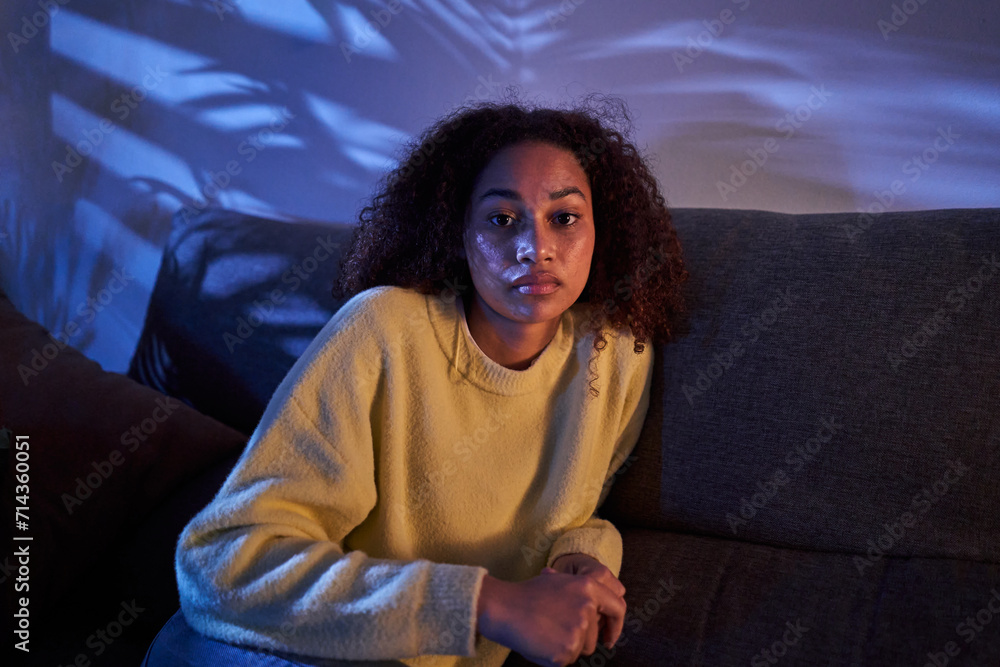 Fototapeta premium Portrait young African American woman looking serious on camera sitting alone on sofa at night home. Afro female posing with sad expression indoors. Photo of lonely, worried and dissociated black girl