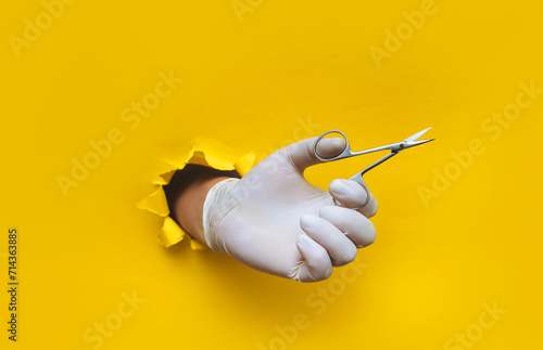 A man's hand in a white medical glove holds a nipper (scissors) for a pedicure and manicure.Yellow paper background with a torn hole in yellow paper.The concept of a master nails of people and animals photo