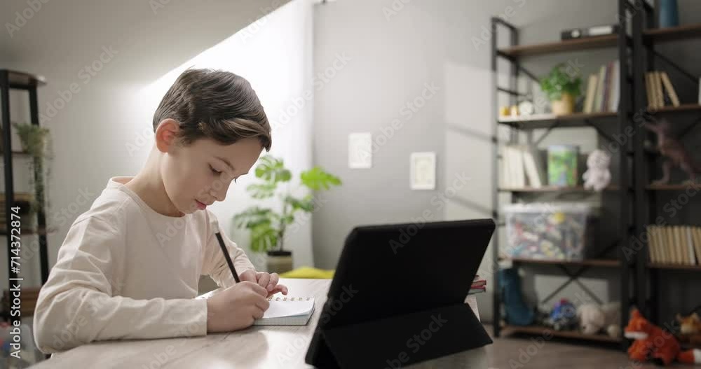 Cute boy studying online from home talking with tutor teacher by video call on digital tablet at home, making notes in copybook. Children remote education Cute boy studying online from home talking with tutor teacher by video call on digital tablet at home, making notes in copybook. Children remote education
