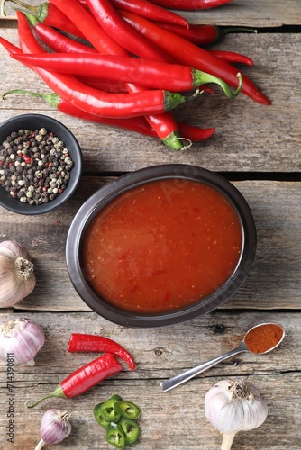 Spicy chili sauce in bowl and ingredients on wooden table, flat lay