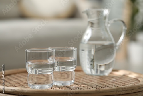 Jug and glasses with clear water on wicker surface against blurred background, closeup