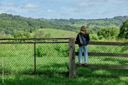 A young girl in blue denim jeans and jacket stands on a fence looking out into the fields and hills.