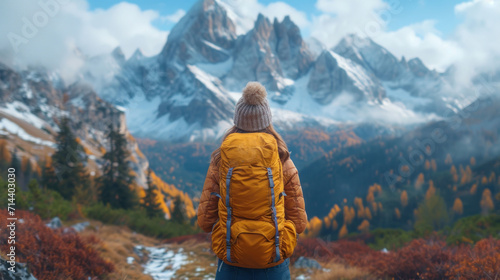  a person in a yellow jacket is looking at a mountain range with snow on the top and trees with orange leaves in the foreground and a blue sky with white clouds.