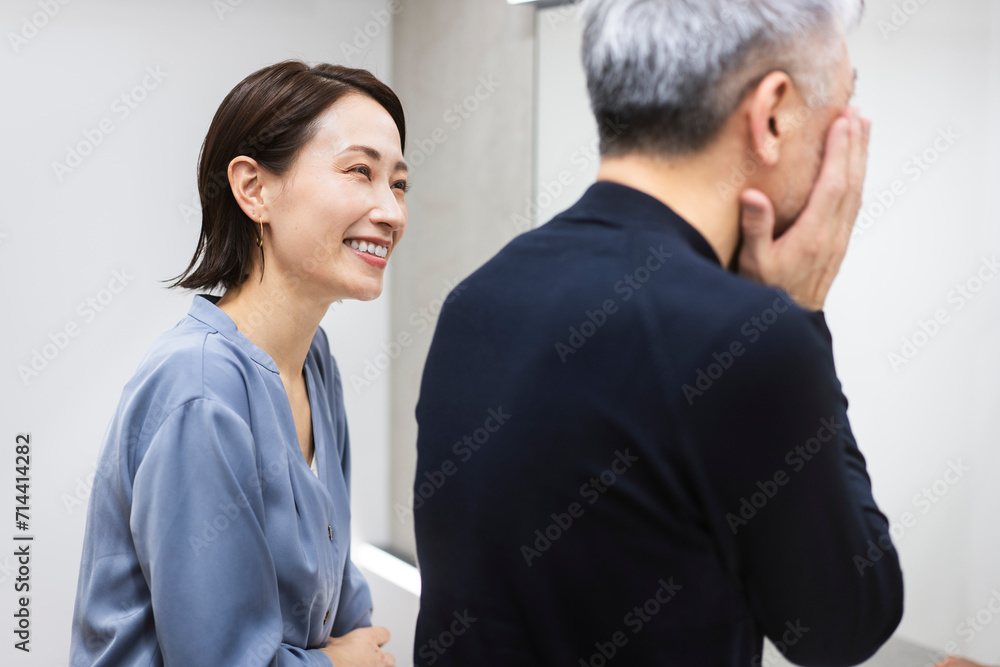 鏡を見ながらスキンケアをしているミドル世代の男性/女性がスキンケアを教えている/メンズビューティー
