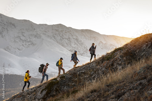 Group of young active hikers with backpacks walks uphill in mountains at sunset