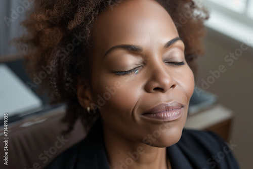  Black businesswoman, eyes closed, leans back in thought. Minimalist office, laptop, notebook - quiet power captured in this close-up portrait. Ample copy space. © Dinusha
