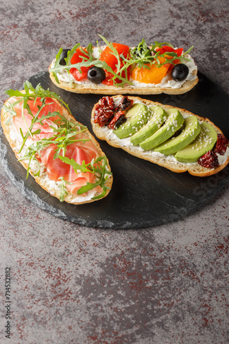 Three different open sandwiches with arugula, jamon, sun-dried tomatoes, avocado, cream cheese, olives and roasted peppers close-up on a slate board on the table. Vertical