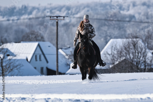 A young red-haired woman rides her horse at a walk across a Schebeckte meadow with a snowy village in the background on a sunny cold winter day.