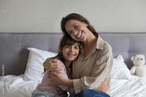 Cheerful young mother hugs cute little daughter sitting on bed in bedroom, enjoy weekend at home, beautiful family looking at camera, pose, showing love, support and care. Happy motherhood, childcare