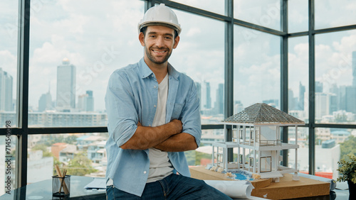 Portrait of architect engineer in casual outfit smile at camera while crossing arms. Engineer looking at camera and sitting with arms folded near house model while wearing safety helmet. Tracery photo