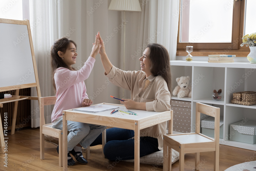 Hispanic mother and little girl sit at table in nursery giving high five gesture, parent praising daughter for well-done assignment, drawing together with pencils in sketchbook. Family pastime, hobby Hispanic mother and little girl sit at table in nursery giving high five gesture, parent praising daughter for well-done assignment, drawing together with pencils in sketchbook. Family pastime, hobby