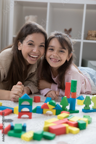 Vertical shot happy young woman lying on warm floor in cozy playroom with little adorable daughter laughing, look at camera enjoy playtime using multicolored wooden blocks set, spend leisure at home