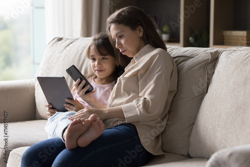 Young woman and 5s daughter using cellphone and tablet at home Little cute girl holding pad device spend time on internet with mother showing her new mobile app, having fun online. Modern tech overuse