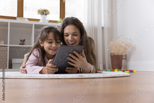 Pretty young Latina woman and daughter spend time on internet lying on warm floor in cozy nursery with digital tablet, watch funny videos or cartoons on-line, buying, make order enjoy new application