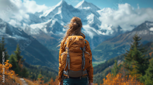  a woman with a backpack standing on a mountain trail looking at a mountain range with a snow capped peak in the distance with trees and yellow flowers in the foreground.