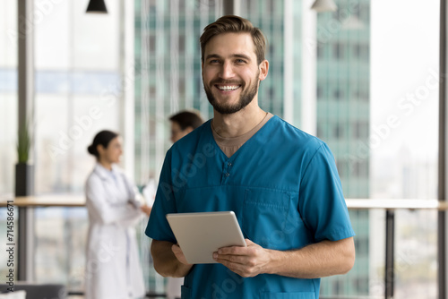 Cheerful handsome surgeon doctor man in blue uniform holding digital tablet computer, looking at camera, smiling, posing for portrait in clinic hall, promoting modern technology in medical job