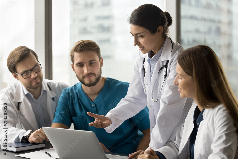 © fizkes - Young Indian doctor woman showing online service for practitioners, speaking at laptop. Multiethnic medical colleagues discussing healthcare application, looking at computer screen at workplace
