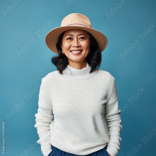 Happy middle aged asian woman in a stylish panama hat and a white sweater. Portrait of senior Japanese woman in a summer hat smiling on a blue background looking at the camera. Cheerful woman in a hat