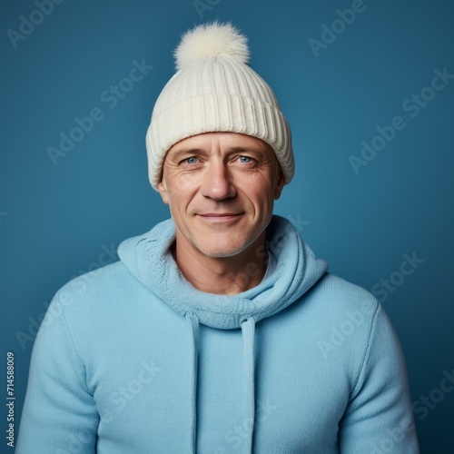 Happy middle aged man in a white winter hat and a  blue sweatshirt. Portrait of a senior man in a winter cap standing on a blue background looking at the camera. Cheerful mature man in a pom pom hat.