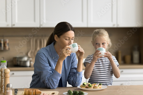 Beautiful young mom and little preschool kid having breakfast at home, sitting at kitchen table, drinking hot beverage, eating sandwiches with fresh vegetables. Mother feeding child photo