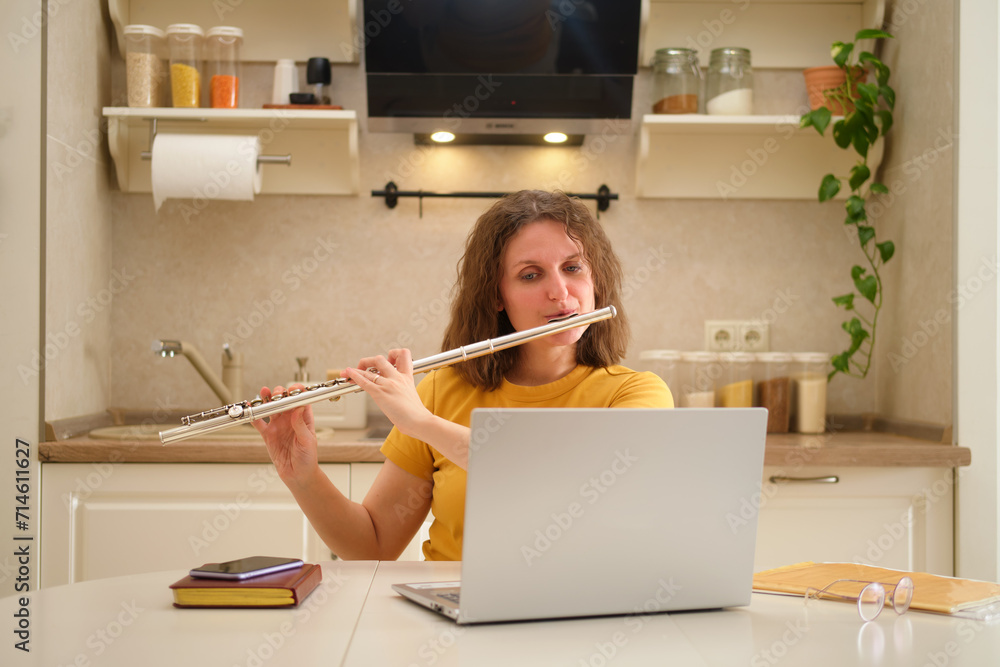 A woman with a laptop playing a flute is sitting at a table in a home kitchen. An adult female businesswoman works from home, a remote office A woman with a laptop playing a flute is sitting at a table in a home kitchen. An adult female businesswoman works from home, a remote office