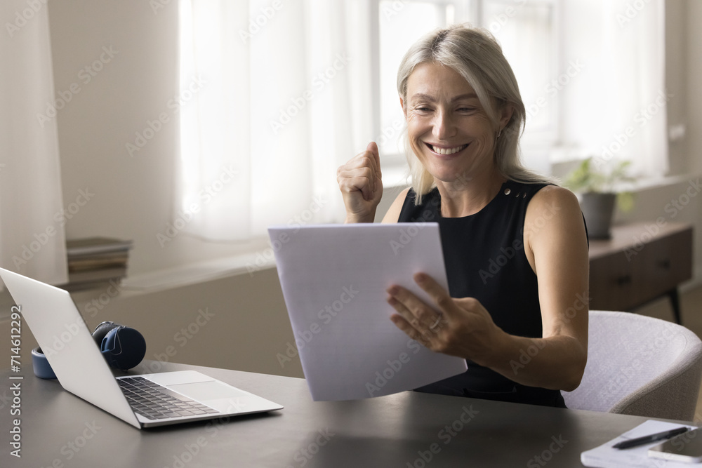 Happy attractive middle aged woman sit at table read postal letter correspondence, get good news in paper notice, make yes gesture, feels happy, receive loan approval, taxes refund, celebrate success Happy attractive middle aged woman sit at table read postal letter correspondence, get good news in paper notice, make yes gesture, feels happy, receive loan approval, taxes refund, celebrate success