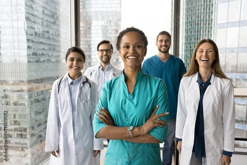 Positive beautiful young African American medical employee woman in blue uniform posing for professional portrait with positive team of doctors behind, looking at camera with hands crossed photo