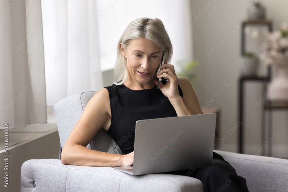 Fototapeta premium Middle-aged woman sit on armchair lead formal conversation, buy on-line, make order using laptop. Electronic commerce client receive consultation by phonecall hold smartphone spend time at home. Tech