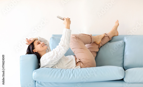 Young brunette woman laying down on cozy sofa in relax position with mobile phone in hands