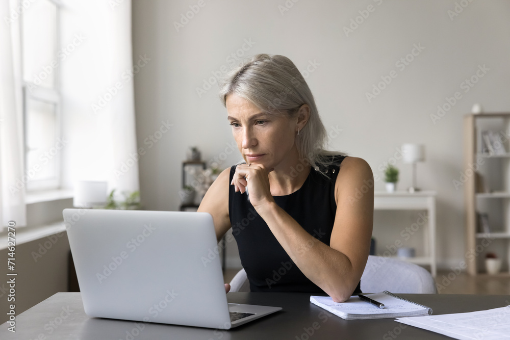 Serious mature businesswoman learn professional program, working on-line, do telecommute freelance remote job, looks at laptop screen seated at desk in homeoffice. Lack of understanding, new software Serious mature businesswoman learn professional program, working on-line, do telecommute freelance remote job, looks at laptop screen seated at desk in homeoffice. Lack of understanding, new software