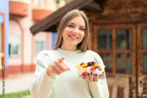 Young pretty blonde woman holding a bowl of fruit at outdoors points finger at you with a confident expression