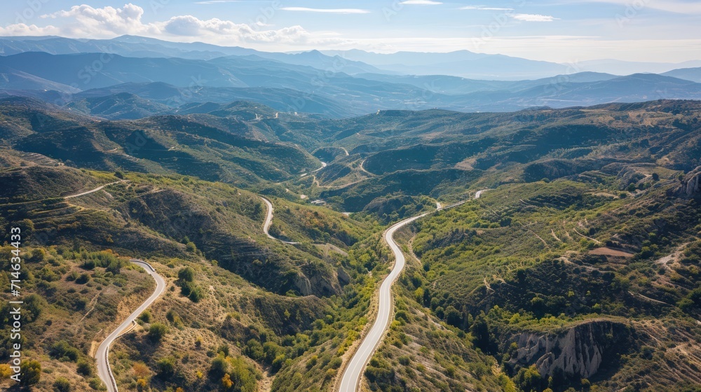 an aerial view of a winding road in the mountains with a view of a valley and mountains in the distance with a blue sky and white clouds in the background. an aerial view of a winding road in the mountains with a view of a valley and mountains in the distance with a blue sky and white clouds in the background.