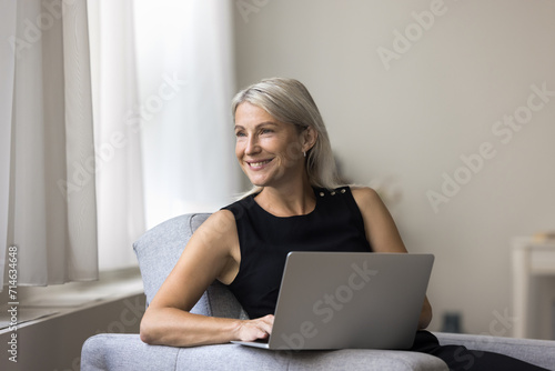 Dreaming mature woman relax at home alone with laptop, sit on armchair looks away, thinking, enjoy online communication with friends, ponders answer, positive freelancer dream about new opportunities