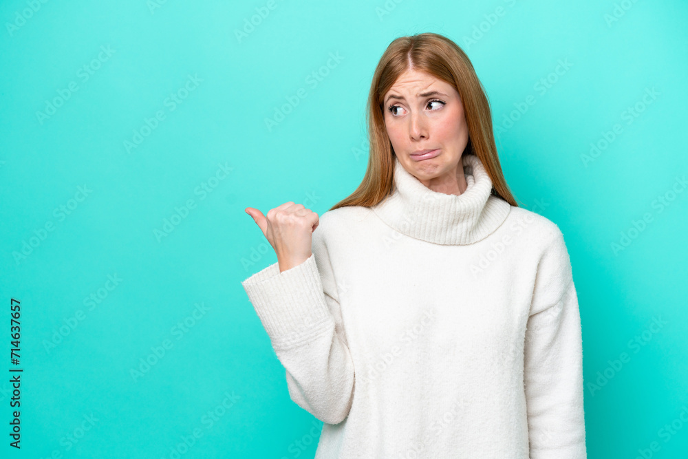 Young redhead woman isolated on blue background unhappy and pointing to the side Young redhead woman isolated on blue background unhappy and pointing to the side