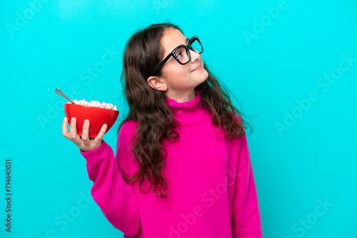 Little girl holding a bowl of cereals isolated on blue background looking up while smiling photo