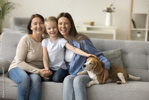 Happy sweet preschool kid girl stroking cute beagle dog on couch  sitting with loving mom and grandma  looking at camera  smiling. Cheerful pretty girls and women of three family generation portrait