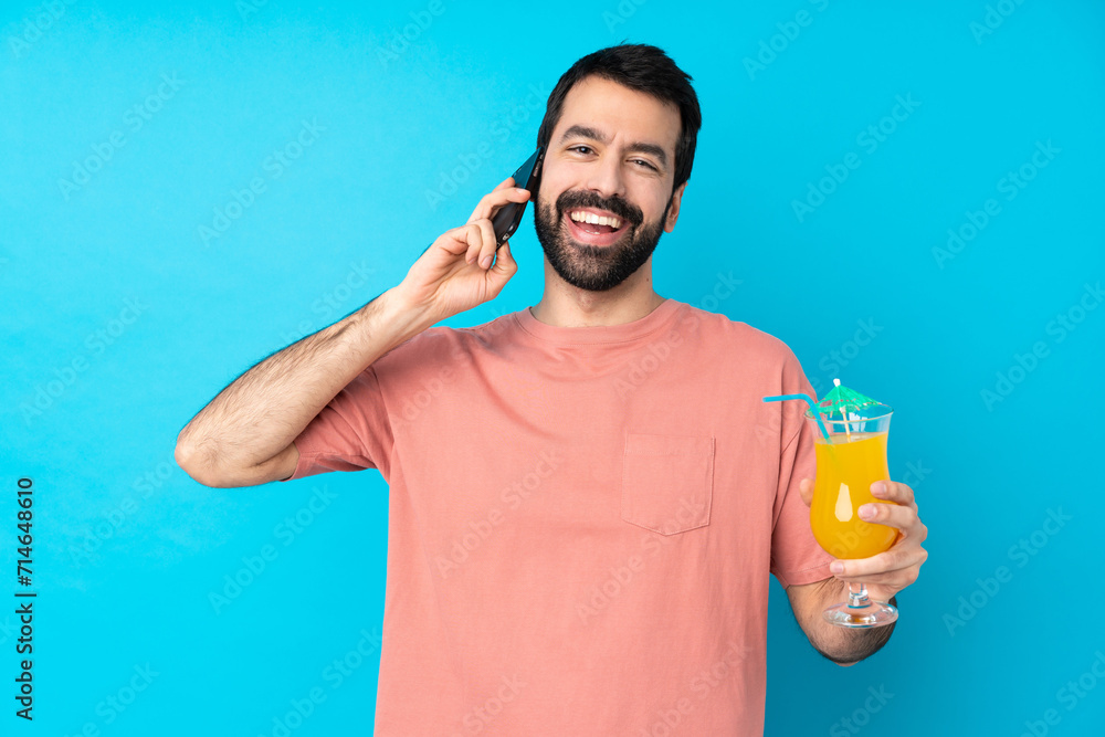 Young man over holding a cocktail over isolated blue background keeping a conversation with the mobile phone with someone Young man over holding a cocktail over isolated blue background keeping a conversation with the mobile phone with someone