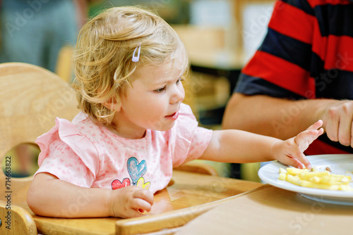 Toddler girl eating healthy vegetables and unhealthy french fries potatoes. Cute happy baby child taking food from parents dish in restaurant. Father eating in fast food restaurant with daughter