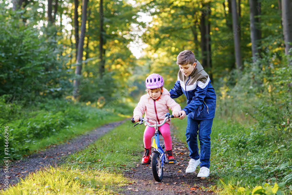 Cute little preschool girl in safety helmet riding bicycle. School kid boy, brother teaching happy healthy sister child cycling and having fun with learning bike. Active siblings family outdoors. Cute little preschool girl in safety helmet riding bicycle. School kid boy, brother teaching happy healthy sister child cycling and having fun with learning bike. Active siblings family outdoors.