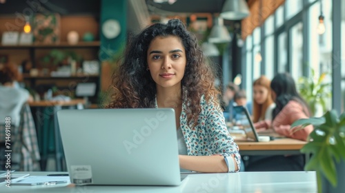 Beautiful Middle Eastern Manager Sitting at a Desk in Creative Office. Young Stylish Female with Curly Hair Using Laptop Computer in Marketing Agency. Colleagues Working in the Background  © Hope