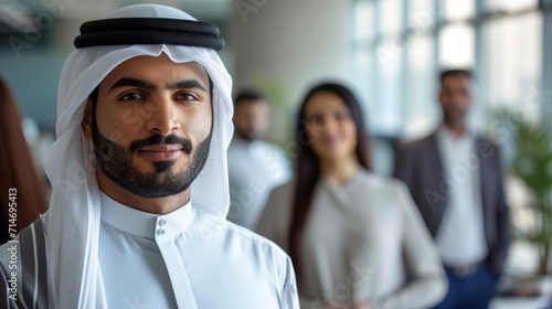 Emirati businessman at office wearing kandura looking at front ideal for middle east business concept. Arabic man inside a corporate establishment with colleagues at the background.  photo