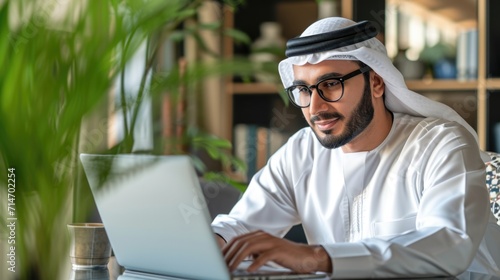 Successful Arab Businessman in Glasses and White Traditional Outfit Sitting in Office and Working on Laptop. Business Manager Planning Corporate Strategy. Saudi, Emirati, Arab Businessman Concept.
