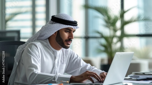 Successful Arab Businessman in White Traditional Outfit Sitting in Office and Working on Laptop Computer. Business Manager Planning Corporate Strategy. Saudi, Emirati, Arab Businessman Concept.